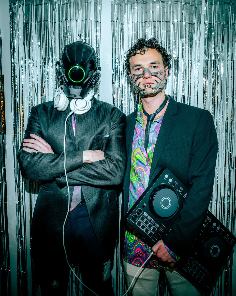 Two DJs in masks with a controller, posing against silver tinsel